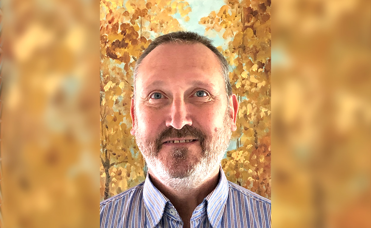Headshot of Head of Business Continuity, Robert West, wearing a light blue pin-striped shirt and standing in front of a painting of autumnal trees