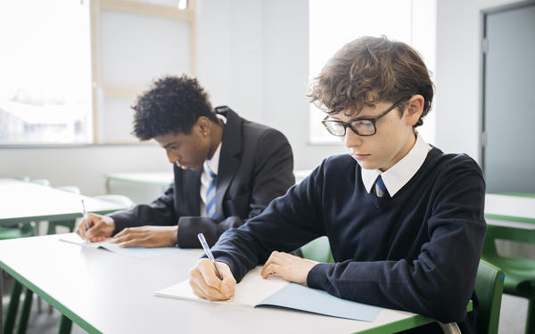 Waist up view of 15 year old secondary schoolboys in uniform, sitting side by side at classroom desk, and writing in papers.