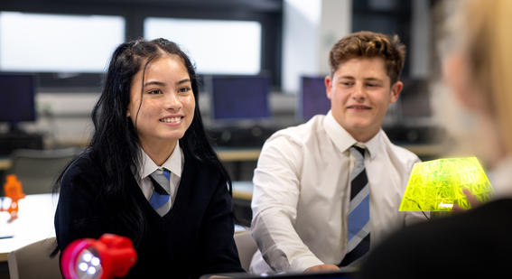 An over-the-shoulder view of two teenage students in their school uniforms having a conversation in the classroom during a lesson in a class in school in the North East of England.